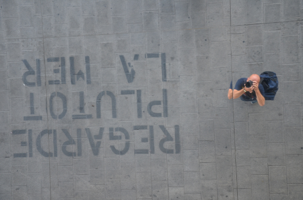 Ein Mann in einem blauen Hemd und Rucksack hält eine Kamera und macht Fotos, wobei Text auf einer Wand oder einem Gehweg links von ihm zu sehen ist.