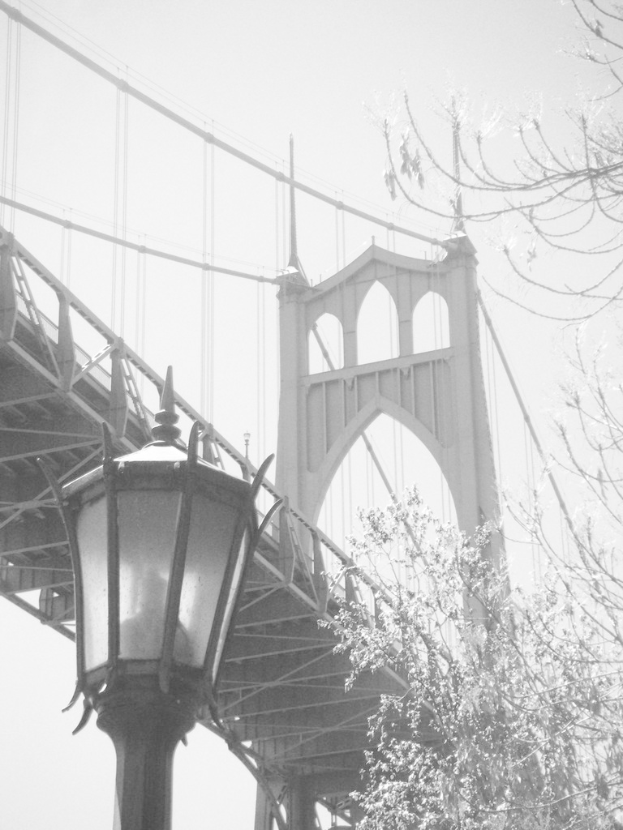 Ein altes Bild einer H├Ąngeseilbr├╝cke mit einem schwarzen Laternenmast vorne unten und einem Baum in der rechten Ecke.