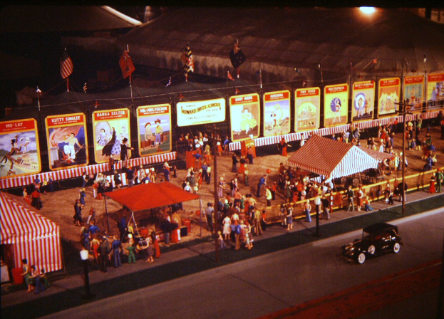 Eine alte nächtliche Faschingszene mit Menschen, einem Auto, Pfählen mit Fahnen, einem Zaun, einem Zelt, Brettern mit Bildern und Text sowie einer Straßenlaterne.