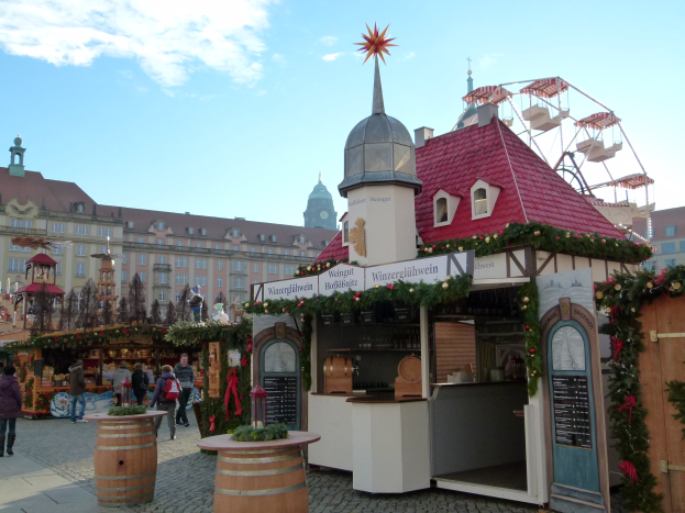 Ein geschäftiger Weihnachtsmarkt in Nürnberg, Deutschland mit Menschen um dekorierte Stände, festlicher Beleuchtung, Schmuck, einem Riesenrad im Hintergrund, Gebäuden mit Fenstern und einem bewölkten Himmel.