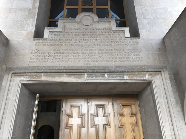 Große hölzerne Tür vor einem steinernen Gebäude mit einem Glasfenster oben, das likely eine Inschrift auf der Wand trägt, die Holocaust-Opfer gedenkt.