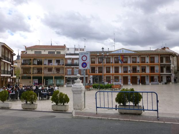 Ein belebter Stadtplatz mit Menschen auf Stühlen sitzend und stehend, umgeben von Topfpflanzen, Metallabsperrungen, Straßenmasten mit Flaggen, ein Schild, Gebäude mit Fenstern und einem bewölkten Himmel.