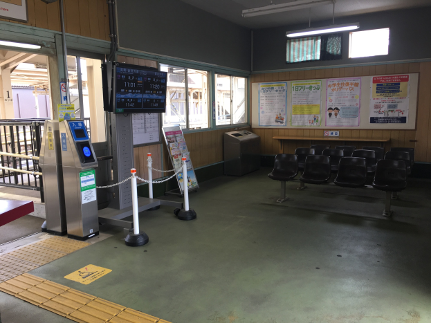 Ein belebter Bahnhofseingang mit Sitzgelegenheiten, einem Tresen, Ticketautomaten, Pfosten mit Ketten, Informationsanzeigen, Plakaten, Deckenleuchten und Geländern.