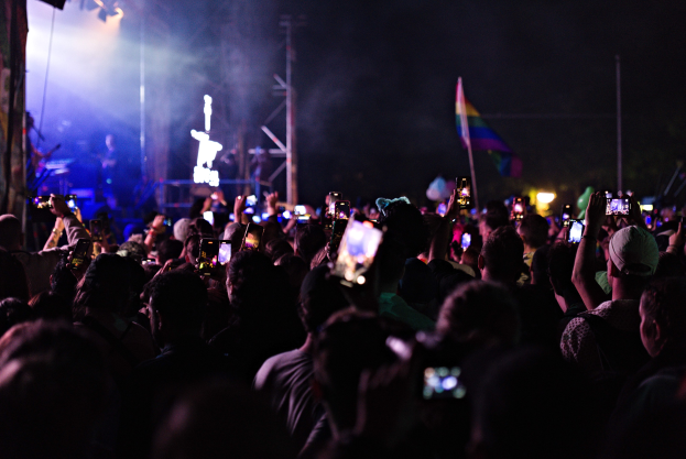 Eine Menge bei einem Konzert mit hochgehaltenen Handys, beleuchtet von Scheinwerfern, umgeben von Pfählen und einer Fahne, während Musiker auf der Bühne spielen.