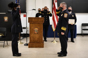 Eine Gruppe uniformierter und maskierter Personen steht vor einem Podium mit Mikrofon, umgeben von Fahnen, Stühlen und Tischen, während eine neue Feuerwehrchef vereidigt wird, wobei eine Person ein Zertifikat hält.