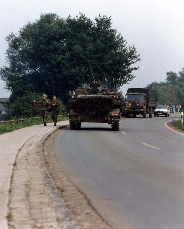 Ein militärisches Fahrzeug fährt eine Straße neben einem Wald entlang, während uniformierte und helmtrugende Individuals neben ihm hergehen, während im Hintergrund Gebäude und ein klarer blauer Himmel zu sehen sind.