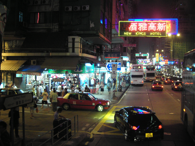 Eine belebte Stadtstraße bei Nacht mit Fahrzeugen auf der Straße, Fußgängern auf dem Gehweg und beleuchteten Gebäuden im Hintergrund unter einem dunklen Himmel.