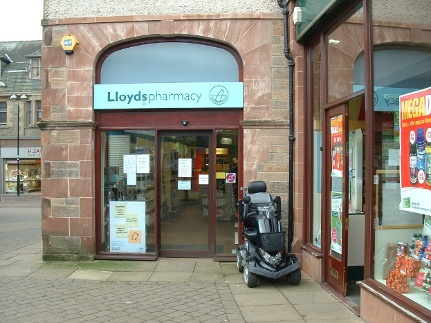 Eine Apotheke mit einem vor ihr geparkten Fahrzeug und einem im linken Eck sichtbaren Gebäude.