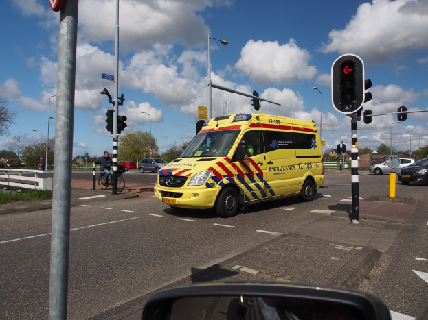 Ambulanz fährt auf einer Straße neben einer Ampel, mit einem Radfahrer im Vordergrund, Laternenpfählen, Bäumen, Gebäuden und einem bewölkten Himmel im Hintergrund und einem Spiegel unten.