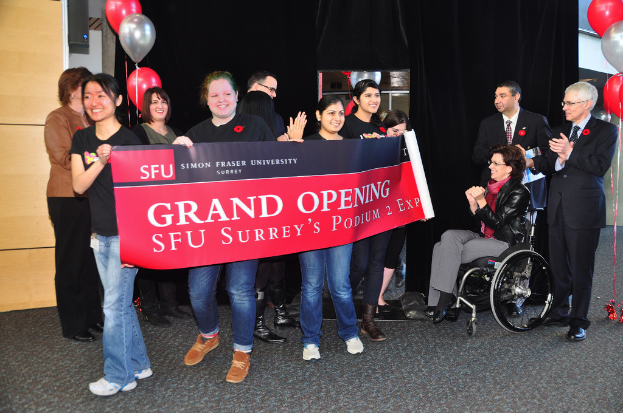 Eine Gruppe von Frauen hält ein Banner, um ein Ereignis zu bewerben, mit zwei klatschenden Männern daneben und einer Frau im Rollstuhl, die aufgeregt aussieht, vor einem schwarzen Vorhang und Ballons.