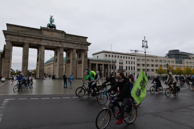 Eine Gruppe von Menschen auf Fahrrädern fährt eine Straße vor dem Brandenburger Tor in Berlin, Deutschland, entlang, wobei einige Taschen tragen, unter einem Himmel mit sichtbaren Laternen und Bäumen.