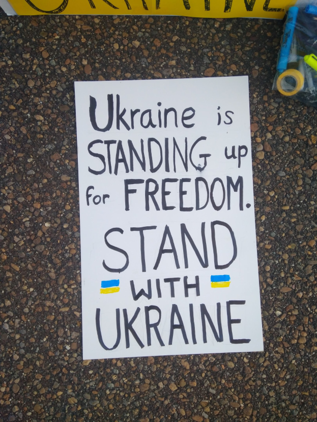 Ein weißes Papier mit den Worten "Ukraine steht für Freiheit auf, steht mit Ukraine" liegt vor einer gelben Fahne auf dem Boden. Auf der rechten Seite befindet sich eine Plastikhülle mit verschiedenen Gegenständen.