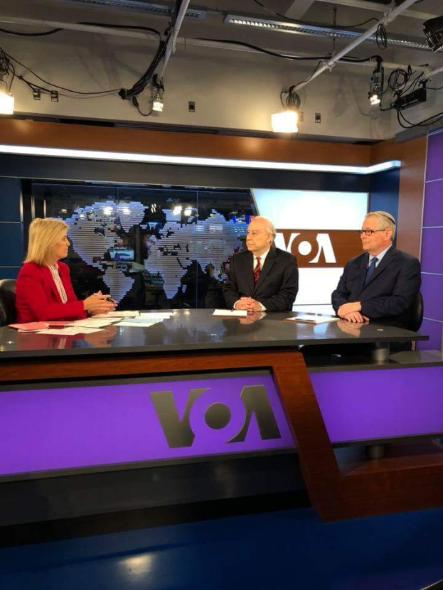 Drei Personen, zwei Männer in Blazern und Krawatten und eine Frau in einem roten Kleid, sitzen an einem Schreibtisch mit Papieren und anderen Gegenständen und führen eine Diskussion vor einem Fernseher mit einem Bildschirm und Lichtern im Hintergrund.