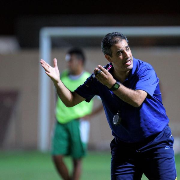 Ein Mann in einem blauen T-Shirt und Hose steht auf einem Fußballfeld und hält ein Mikrofon in der Hand, während er gestikuliert, als würde er trainieren. Im Hintergrund sind ein Torpfosten und eine Person in einem grünen Kleid zu sehen.