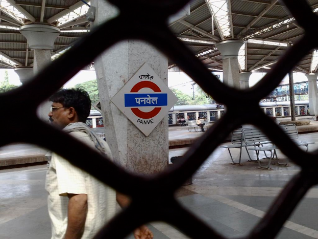 Ein Mann steht auf einem Podest hinter einer Maschendrahtbarriere, mit Bänken und Säulen in der Nähe, unter einem Dach, mit einem Zug, einer Brücke und Bäumen im Hintergrund.