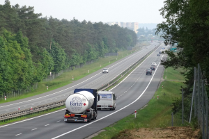 Ein großer Lkw fährt auf einer von Bäumen gesäumten Autobahn, mit einem Zaun und Gras auf der rechten Seite und Gebäuden in der Ferne unter einem klaren blauen Himmel.