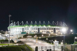 Ein großes Stadion bei Nacht beleuchtet mit parkenden Autos davor, umgeben von Straßenlaternen, Strommasten, Stromkabeln, Fahrzeugen auf der Straße, Gittern, Bäumen und einem sternenklaren Himmel.