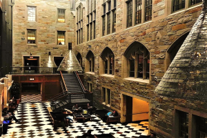 Großer Raum mit schwarz-weißem Schachbrettboden, zentraler Treppe, Geländern, Fenstern, Deckenleuchten und mehreren Personen an Tischen und Stühlen sitzend, Teil einer Harry-Potter-Studiotour in London.