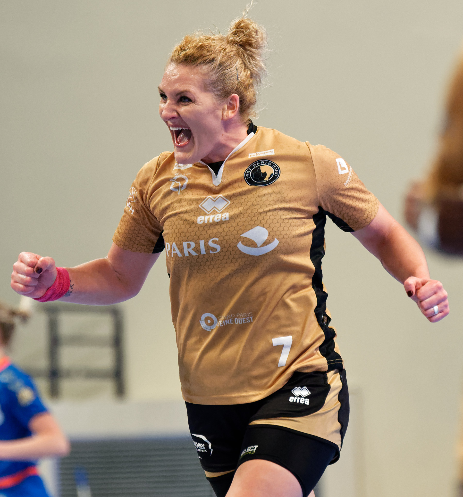 Eine Frau in einem braunen Hemd und schwarzen Shorts rennt auf einem Futsalplatz, feiert mit einem triumphalen Ausdruck, während zwei andere Frauen im Hintergrund unscharf sind.