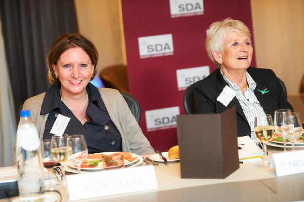 Zwei Frauen sitzen an einem Tisch mit Essen, Getränken und verschiedenen Gegenständen, sie lächeln, mit einem Banner und Vorhängen im Hintergrund.