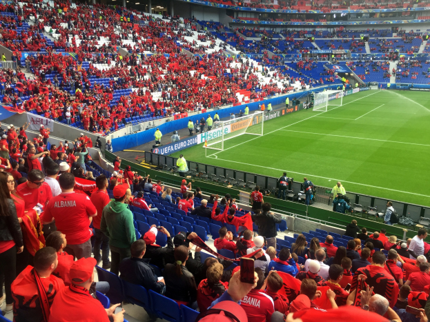 Eine große Menschenmenge in einem hell erleuchteten Stadion schaut einem Fußballspiel zu, mit Menschen, die auf Stühlen sitzen und stehen, Fahnen, einem Torpfosten und Deckenbeleuchtung.