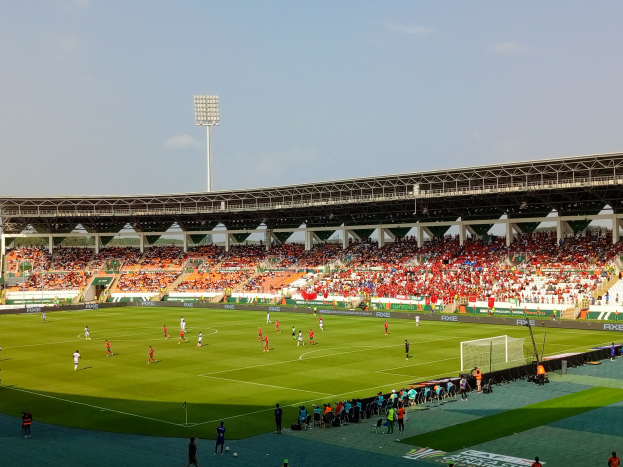 Ein Fußballspiel in einem großen Stadion mit Spielern auf dem Boden, einem Tor, einem Zaun, einer Fahne mit Text, Metallrahmen, einem Dach mit Deckenlampen und einem bewölkten Himmel.
