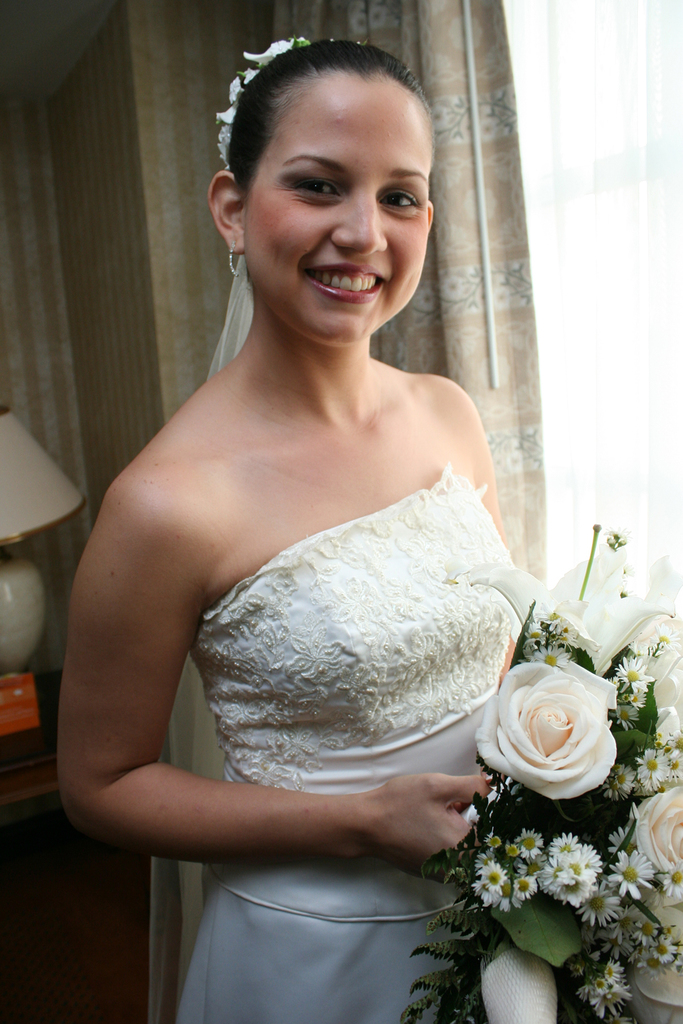 Eine Braut steht strahlend in der Mitte und hält einen Blumenstrauß, mit einem Vorhang und einer Wand im Hintergrund und einer Lampe auf einem Tisch links.