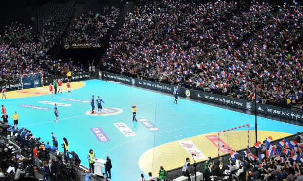 Eine Gruppe von Menschen, die Handball in einem Stadion mit einem Netz auf dem Feld und Zuschauern in den Rängen spielen, während Fotografen die Aktion aufnehmen.