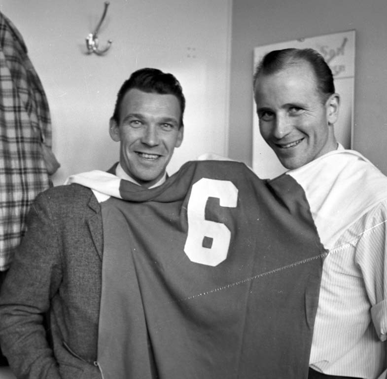 Zwei Männer in Schwarz und Weiß, die strahlend ein Fußballtrikot hochhalten, mit einer Tafel und einem Bügel an der Wand dahinter.