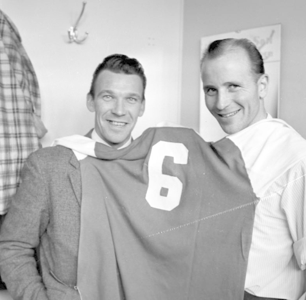 Zwei Männer in Schwarz und Weiß, die strahlend ein Fußballtrikot hochhalten, mit einer Tafel und einem Bügel an der Wand dahinter.