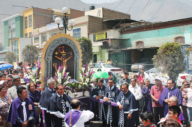Menschen auf einer Straße mit einem Kreuz und Blumensträußen in der Mitte, Autos dahinter und Gebäude, Bäume und Straßenlaternen im Hintergrund.