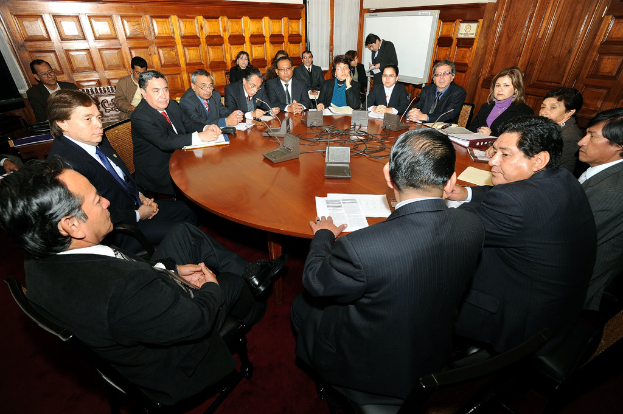 Eine Runde-Tisch-Besprechung mit Delegierten, die über Mikrofone, Papiere, Kabel und Dateien auf dem Tisch und eine hölzerne Wand im Hintergrund diskutieren.