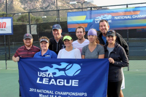 Gruppe von Menschen auf einem Tennisplatz mit einem "2015 USTA National Championships"-Banner, einem Zaun, Bannern, Laternenmasten, B├Ąumen, Bergen und einem klaren blauen Himmel im Hintergrund.