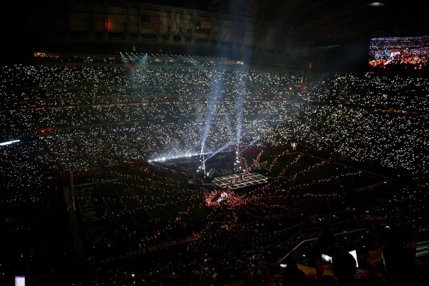 Eine große Menge in einem hell erleuchteten Stadion bei Nacht schaut auf einen Bildschirm, der die Ergebnisse der Super Bowl XLVII-Halbzeitshow anzeigt.