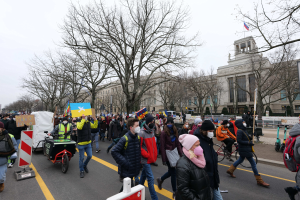 Eine große Protestdemonstration mit Menschen, die eine Straße in Washington, D.C. entlanggehen, einige mit Schildern und Fahrrädern, unter einem klaren blauen Himmel mit Bäumen und Hinweistafeln im Hintergrund.