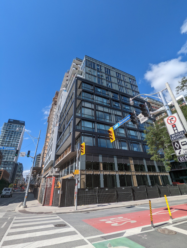 Eine belebte Stadtstraße mit einem hohen Apartment/Wohnhausgebäude zum Verkauf an 541 East 57th Street, Unit 541, Verkehrszeichen, Fahrzeuge, Schilder, Bäume, ein Zaun und bewölkter Himmel.