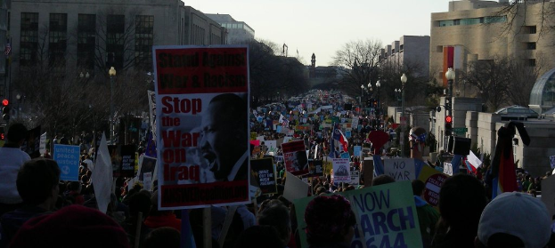Menschen protestieren auf einer Stra├če und halten Plakate, mit Geb├Ąuden, B├Ąumen, Laternen und dem Himmel im Hintergrund.