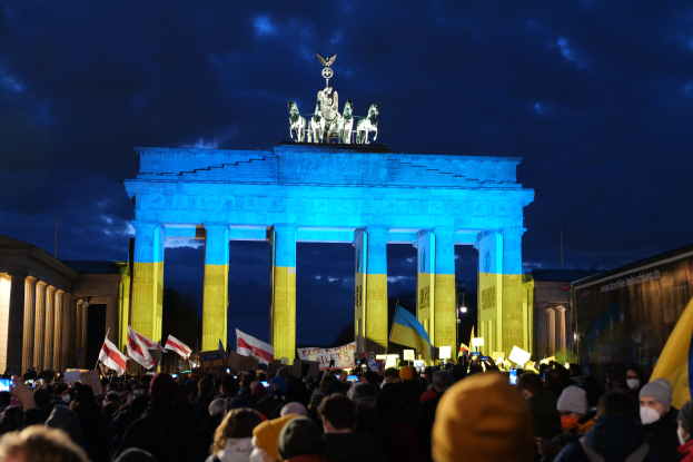 Eine Menschenmenge steht vor dem Brandenburger Tor in Berlin, Deutschland, mit Fahnen und Plakaten in den Händen, mit einem Banner auf der rechten Seite und dem Tor mit Statuen und Säulen geschmückt unter einem bewölkten Himmel.