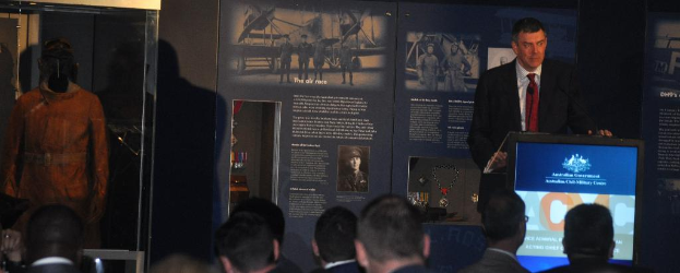 Eine Person steht an einem Podium mit einer Gruppe von Menschen davor, eine Puppe in einer Kiste links daneben und Tafeln im Hintergrund mit Text, Medaillen und Bildern.