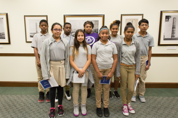 Eine Gruppe junger Schüler mit Büchern vor einer Wand mit Fotorahmen, was auf eine Schulumgebung hinweist.