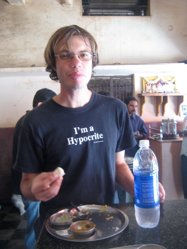 Eine Person sitzt an einem Tisch in einem Hotel, hält Essen in ihrer rechten Hand und hat eine Flasche auf dem Tisch, während im Hintergrund andere Menschen, ein Gotteskultbild, ein Fenster und eine Wand zu sehen sind.