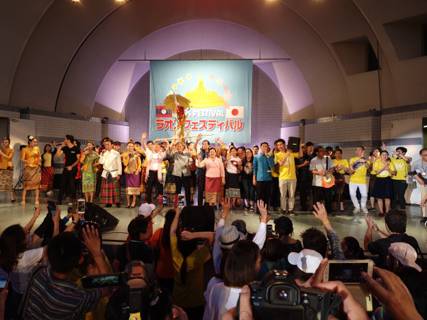 Eine Gruppe von Menschen auf einer Bühne mit Kameras, Handys und Musikinstrumenten während einer scheinbaren Festival-Eröffnung, mit einem Banner, Wänden und Deckenlampen im Hintergrund.