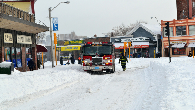 Ein Feuerwehrauto steht im Schnee vor Gebäuden, mit Geschäften und Laternenmasten links, einer Ampel in der Mitte und Schnee auf dem Boden.