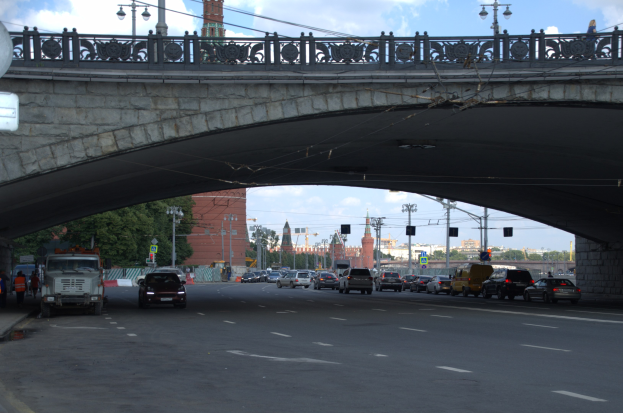 Eine Brücke spannt eine vielbefahrene Straße, unter der Autos und Lastwagen fahren, während Fußgänger auf dem linken Gehweg stehen, mit städtischen Elementen wie Ampeln, Laternen, Bäumen, Gebäuden und Himmel im Hintergrund.