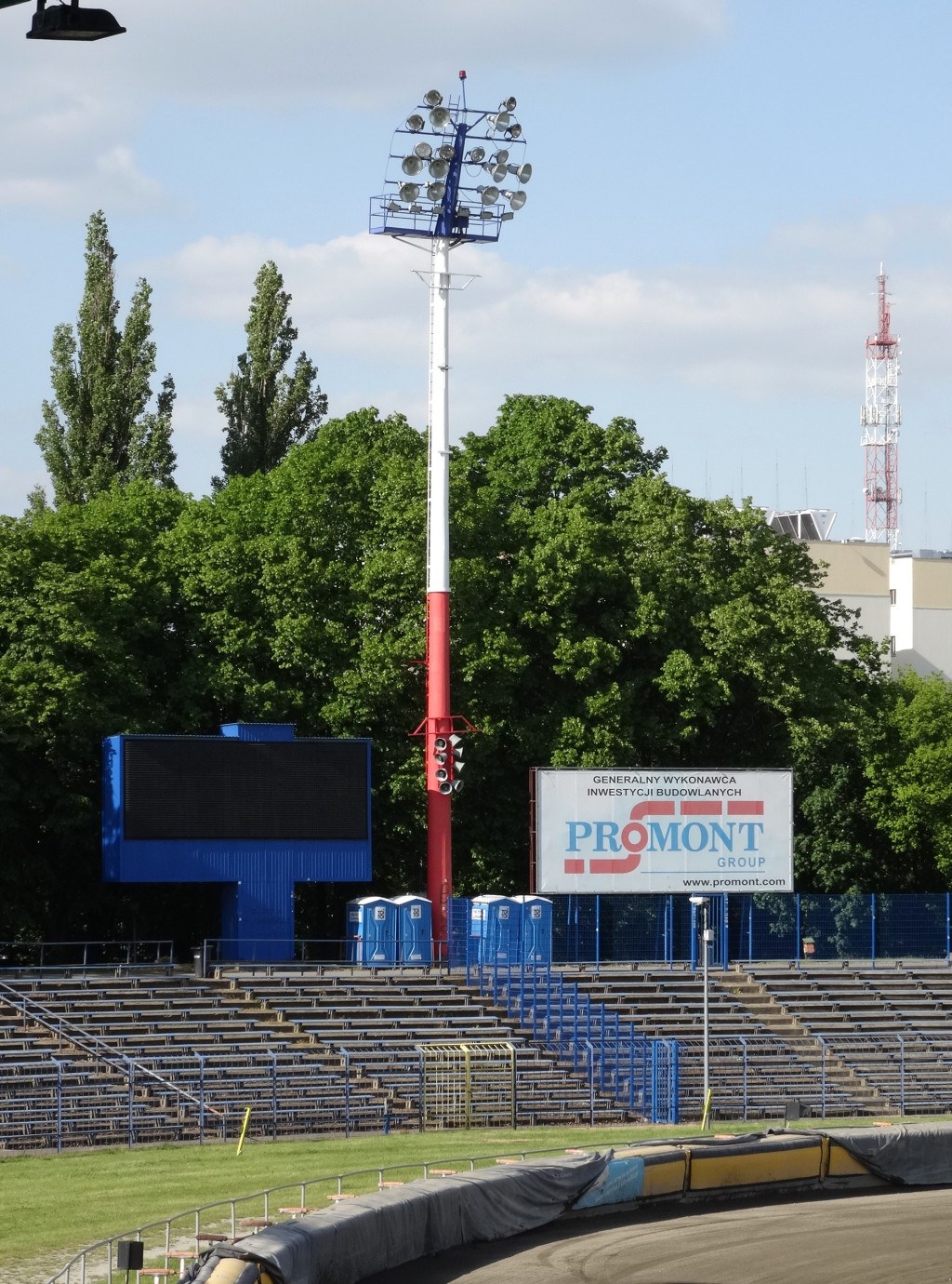 Rennstrecke beleuchtet von oben, umgeben von Gras, Geländern, Stufen, einem Lichtmast, einem Schild, Bäumen, Gebäuden, Türmen und einem bewölkten Himmel.