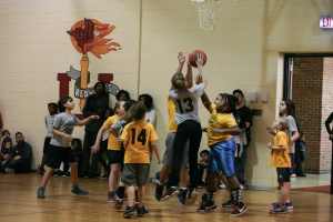 Kinder in gelben und grauen T-Shirts spielen Basketball, wobei ein Mädchen in einem grauen T-Shirt mit der Nummer 13 den Ball wirft. Ein Mann in einem blauen Hemd sitzt auf der linken Seite des Courts, mit einer weißen Wand mit Text dahinter. Rechts ist ein Ausgangsschild und ein rotes Backsteingebäude zu sehen.