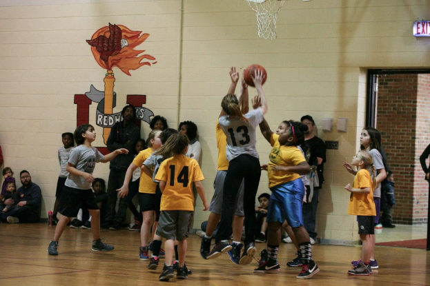 Kinder in gelben und grauen T-Shirts spielen Basketball, wobei ein Mädchen in einem grauen T-Shirt mit der Nummer 13 den Ball wirft. Ein Mann in einem blauen Hemd sitzt auf der linken Seite des Courts, mit einer weißen Wand mit Text dahinter. Rechts ist ein Ausgangsschild und ein rotes Backsteingebäude zu sehen.
