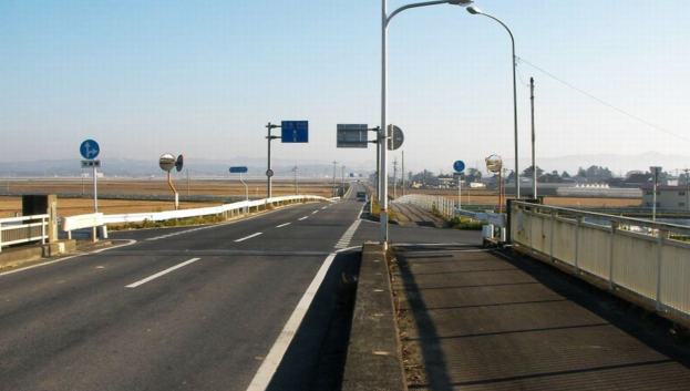 Autobahn mit einer Brücke darüber, mit Geländern, Laternenmasten und Verkehrszeichen; Gebäude, Bäume, Hügel und einen klaren blauen Himmel im Hintergrund.