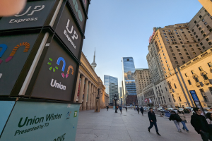 Eine belebte Stadtstraße mit Fußgängern, hohen Gebäuden, Laternen, Fahrzeugen und Texttafeln auf der linken Seite, die sich auf die Union Winter Conference beziehen, sowie einem sichtbaren Himmel im Hintergrund.