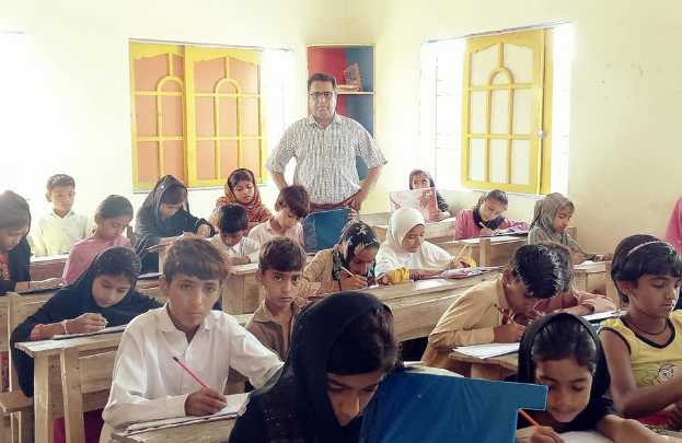 Eine Gruppe von Kindern sitzt an Tischen in einem Klassenzimmer in Pakistan, mit Büchern und Stiften in den Händen, mit einer Erwachsenen im Hintergrund und einem Bücherregal im Hintergrund und Fenstern an den Wänden.